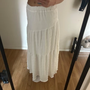 Vit långkjol S/XS - Säljer en superfin vit långkjol med resår i midjan. Kjolen har en luftig och flowy känsla, perfekt för varma dagar. Materialet är lätt och har en snygg struktur som ger kjolen extra liv. 