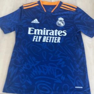 Real Madrid bortatröja Adidas blå 164 - Säljer en blå Real Madrid bortatröja från Adidas i storlek 164. Tröjan har korta ärmar, orange detaljer på axlar och krage, samt ett unikt grafitti-inspirerat mönster. Tillverkad i Primegreen-material, perfekt för fotbollsplanen eller att bära som supporter. Säljer för 600 men nypris är runt 1000 kan sänka pris!