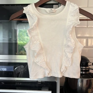 Vit volangtop från H&M - Supersöt vit linnetopp från H&M med breda axelband och volangdetaljer framtill. Toppen är ärmlös och har broderade volanger längs sidorna som ger en extra touch. Perfekt för varma dagar och enkel att matcha med jeans eller kjol.