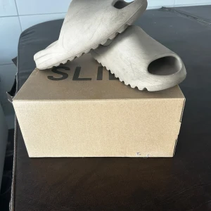 Beige adidas Yeezy Slide sandaler - Säljer ett par adidas Yeezy Slide i beige färg. De har en chunky, modern design med öppen tå och räfflad sula för extra grepp. Tillverkade i syntetmaterial och perfekta för chill dagar eller när du vill ha en clean look. Box medföljer.
