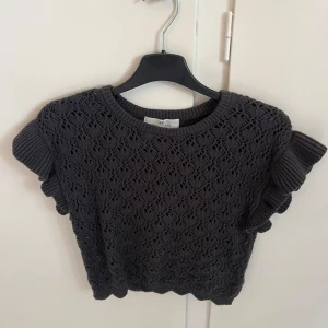 Mörkgrå stickad topp med volangärm - Supersöt mörkgrå stickad topp från H&M med korta volangärmar och mönster över hela plagget. Rund halsringning och ribbade kanter ger en extra snygg touch. Perfekt att styla med jeans eller kjol för en trendig look.