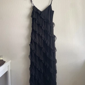 Svart långklänning med volanger - Säljer en svart långklänning med tunna axelband och v-ringning. Klänningen har volangdetaljer och en slit på ena benet