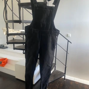 Svarta denim hängslebyxor/snickarbyxor från Monki - Svarta hängslebyxor från Monki i klassisk jeansmodell med raka ben och justerbara axelband. Byxdressen har fickor fram och bak samt knappar i sidorna. Tillverkad i kraftigt bomullsjeans-tyg, perfekt för en chill och avslappnad look.