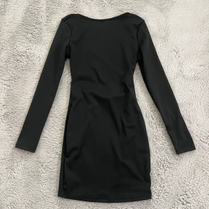 Svart långärmad kortklänning med snörning - Snygg svart kortklänning med långa ärmar och cool snörningsdetalj över ryggen. Klänningen har en figurnära passform och är tillverkad i ett stretchigt material som sitter perfekt. Perfekt för dig som vill ha en stilren men ändå trendig look.