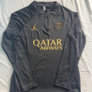 Svart PSG träningströja Jordan med Byxor - Svart långärmad träningströja från PSG x Jordan med guldiga detaljer och loggor. Tröjan har halv dragkedja framtill, diskret mönster i tyget och texten 'Paris' på ärmen. Tillverkad i lätt polyester med Dri-FIT-teknologi för maximal komfort.
