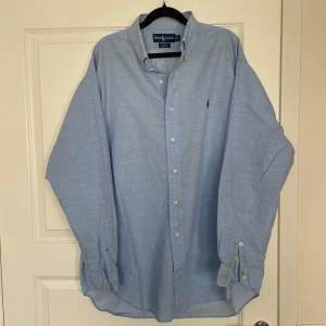 Ljusblå skjorta från Ralph Lauren - Klassisk ljusblå skjorta från Ralph Lauren med button-down krage och broderad logga på bröstet. Skjortan är i 100% bomull och har långa ärmar med knappar vid manschetterna. Perfekt för en clean och stilren look. OBS, har fläck längs ned på ena armen! 