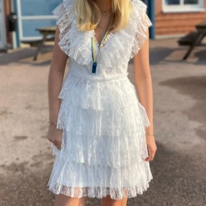 Vit spetsklänning med volanger - Superfin vit kortklänning i spets med flera lager volanger och v-ringning. Klänningen har pärlknappar i ryggen och är ärmlös med bred volangkant. Perfekt för student eller sommarens alla tillfällen. Klänningen är ifrån bubbelroom men jag har varit att ta bort spärrarna där fram, gjort av en skräddare. 