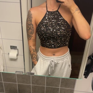 Svart glittrig croppad halterneck topp - Säljer en svart croppad halterneck topp med massor av glittrande paljetter. Toppen knyts både i nacken och ryggen för perfekt passform och har öppen rygg. Perfekt för dig som vill sticka ut och glänsa på festen!