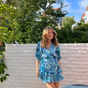 Blå mönstrad volang klänning  Zara - Superfin kort omlottklänning från Zara i blått, turkost och vitt paisleymönster. Klänningen har v-ringning, volangkant nertill och knyt i midjan. Lätt och luftig med korta ärmar, perfekt för varma dagar.