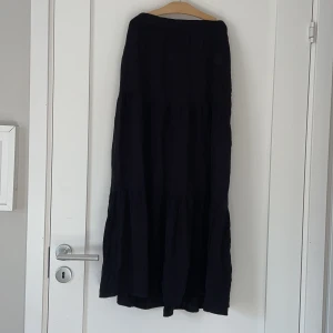 Svart långkjol med knyt i midjan - En svart långkjol med volanger och två snören på varsin sida, man kan antingen knyta där bak eller gömma de i kjolen. Kjolen har en luftig och flowy känsla, perfekt för dig som gillar en avslappnad men ändå stilren look. Passar lika bra till sneakers som boots.