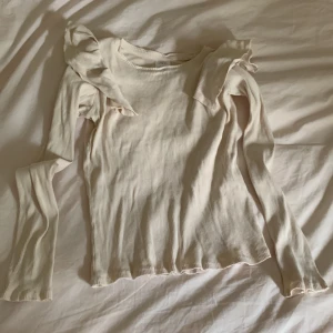 Beige ribbad topp med volang från Zara - Supersöt beige ribbad topp från Zara med långa ärmar och volangdetaljer på axlarna. Figurnära passform och stretchigt material som känns skönt mot huden. Perfekt för dig som gillar en enkel men ändå trendig look.