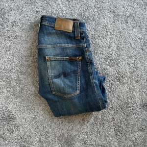 Blå jeans från Nudie Jeans, 27/30 - Snygga blå jeans från Nudie Jeans med klassisk femficksdesign och orange kontrastsömmar. Jeansen har en rak passform och är tillverkade i slitstarkt bomullsdenim. Bakfickan har Nudie Jeans ikoniska broderi. Perfekta för en avslappnad stil.