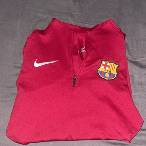 Äkta FC Barcelona Nike långärmad tröja - Säljer en mörkröd långärmad FC Barcelona träningströja från Nike med halv dragkedja. Tröjan har klubbens emblem på bröstet och diskret mönster på ärmarna. Perfekt för dig som älskar fotboll och vill ha en snygg sportig look.