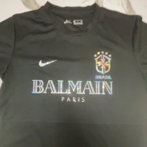 Svart Brasil Nike x Balmain fotbollströja S - Svart fotbollströja från Nike med Brasil-logga och Balmain Paris-tryck i holografisk stil på bröstet. Kortärmad modell i lätt funktionsmaterial (Nike Aeroswift). Snygg och modern look med rund hals och klassisk passform.