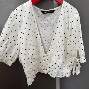 Prickig blus med knyt från Zara - Supersöt vit blus från Zara med svarta prickar, v-ringning och knappar framtill. Blusen har korta ärmar med knyt i sidan och volangkant nedtill. Perfekt för dig som gillar en lekfull och trendig stil.