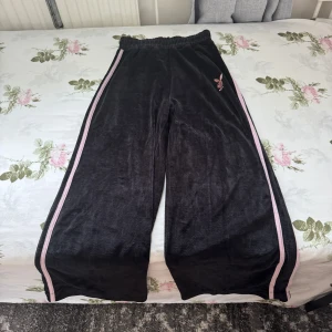 Svarta byxor Playboy x Missguided - Svarta mjukisbyxor från Playboy x Missguided med breda ben, rosa ränder längs sidorna och stor rosa PLAYBOY-logga bak. Resår i midjan och klassisk bunny-logga framtill. Perfekta för en chill och trendig look. 