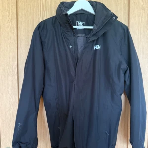 Svart Helly Hansen vinterjacka  - Svart vind- och vinterjacka från Helly Hansen med Helly Tech Protection-tyg. Jackan har quiltat foder, dragkedja och huva. Perfekt för blåsiga dagar och har en clean look med diskret logga på bröstet och ärmen. Snygg och funktionell för dig som gillar outdoor. Använd max 1 gång. 