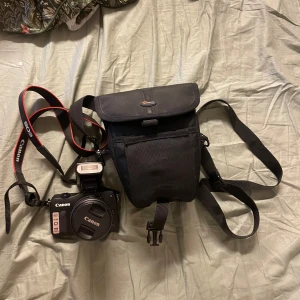 Svart kameraväska från Canon - Laddare fodral följer med kamera! Säljer en svart kameraväska från Lowepro med justerbar axelrem och praktiskt lock med spänne. Väskan är tillverkad i slitstark polyester och har en enkel, stilren design som passar perfekt för att bära systemkamera och tillbehör.