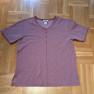 Ljuslila/rosa topp med knappar från Teddi - Kortärmad topp i ljuslila/rosa från Teddi med v-ringning och tre dekorativa knappar framtill. Tillverkad i en mjuk blandning av bomull och polyester. Perfekt för dig som gillar enkel men snygg stil.