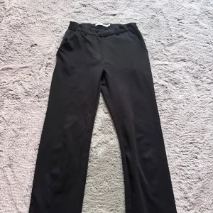 Svarta kostymbyxor från 157 - Klassiska svarta kostymbyxor från 157 med raka ben och midja med hällor för bälte. Byxorna har diskreta sidfickor och stängs med knapp och dragkedja. 