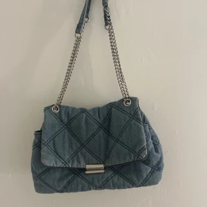 Quiltad jeans axelväska med kedja - Snygg quiltad axelväska i blått jeanstyg med silverfärgad kedjerem och metallspänne. Väskan har lock med rutmönster och praktiskt innerfack med dragkedja. Perfekt för dig som gillar en cool och avslappnad stil. Jag vet inte vilket märke det är.. priset går att diskutera!💕💕