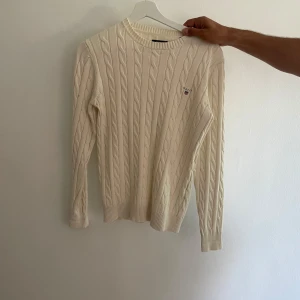 Vit kabelstickad tröja från Gant - Stickad vit tröja från Gant med rund halsringning och diskret logga på bröstet. Tröjan har lång ärm och ribbade muddar vid ärmslut och nederkant. Perfekt för lager-på-lager och en clean look. Mycket bra skick 