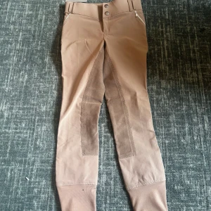 Nya ridbyxor från CRW, stl 160 - Snygga beige ridbyxor från CRW med förstärkta partier på insidan av benen och sätet. Byxorna har dragkedjeficka fram, bälteshällor och tight passform. Perfekta för dig som gillar ridning och vill ha stilrena, funktionella byxor.