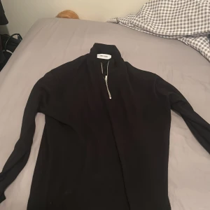 Svart half zip tröja Only & Sons - Svart långärmad tröja från Only & Sons med hög krage och silverfärgad half zip-dragkedja. Tröjan har en clean och stilren look, perfekt för lager-på-lager. Materialet känns mjukt och stretchigt, troligen bomull eller polyester.