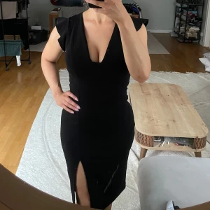 Svart cocktailklänning med vadderade kupor - Mjuk och smickrande festklänning med slits och v-urringning. Vadderade kupor. Mönstrat tyg med knäppning baktill och halva ärmar med volang.