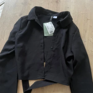 Svart croppad skjorta med knyt bak - Svart croppad skjorta från H&M med långa ärmar och snygg knytning i ryggen. Skjortan har klassisk krage och knappar framtill. Tillverkad i ett mjukt material som känns skönt mot huden. Aldrig använd, lappar kvar.