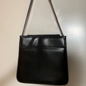 Svart axelväska från ZERA ZERA - Stilren svart axelväska från ZERA ZERA i slätt skinn. Väskan har en fyrkantig form, lång axelrem och minimalistisk design utan synliga detaljer. Perfekt för dig som gillar en clean och klassisk look. I bilderna 3 och 4 ser man lite slitage. Frakt tillkommer