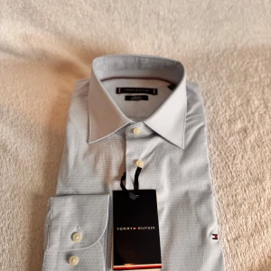Ljusgrå skjorta Tommy Hilfiger Regular Fit - Snygg ljusgrå skjorta från Tommy Hilfiger med diskret mönster och klassisk krage. Regular fit med stretch för extra komfort. Ikonisk liten logga på ärmen och vita knappar framtill. Perfekt för dig som vill ha en stilren look med premiumkänsla.