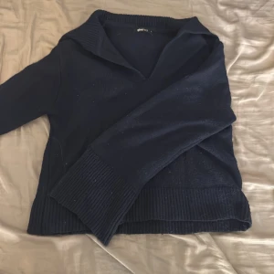 Mörkblå stickad tröja från Gina Tricot - Mysig mörkblå stickad tröja från Gina Tricot med bred ribbad kant vid ärmslut och nederkant. Tröjan har en djup v-ringning och krage som ger en avslappnad men ändå stilren look. Perfekt för lager-på-lager under kyliga dagar.