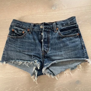 Levi's jeansshorts med fransig kant - Snygga mörkblå jeansshorts från Levi's med fransig, rå kant och klassiska fem fickor. Shortsen har hög midja och knappar framtill. Perfekta för sommaren och ger en cool vintage vibe.