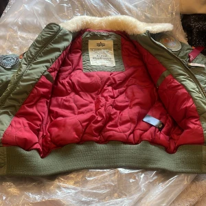 Grön bomberjacka Alpha Industries S - Snygg grön bomberjacka från Alpha Industries med röd quiltad insida, patchar på ärmar och bröst samt vit fuskpäls vid kragen. Jackan har dragkedja, ribbade muddar och flera coola detaljer som ger en riktigt streetig vibe.