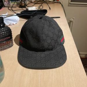  Gucci keps äkta - Svart keps från Gucci med klassiskt GG-monogram över hela kepsen. Sidorna har de ikoniska gröna och röda ränderna. Justerbar rem baktill i svart skinn. Snygg och stilren keps som lyfter vilken outfit som helst.