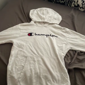 Vit hoodie från Champion med logga - Säljer en vit hoodie från Champion med stor broderad logga över bröstet. Tröjan har huva, magficka och långa ärmar. Perfekt för dig som gillar en clean och sportig stil. Klassisk passform och mjukt material.