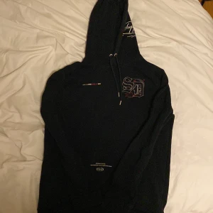 Svart hoodie Supply & Demand NYC - Svart hoodie från Supply & Demand med tryck på bröstet och texten 'Supply' på huvan. Klassisk modell med dragsko i huvan och långa ärmar. Snygga detaljer med grafiskt tryck och brodyr. Perfekt för dig som gillar streetwear och vill ha en stilren look.