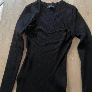 Svart ribbad tröja Luxe To Kill - Svart ribbad klänning från Luxe To Kill med lång ärm och figurnära passform. Klänningen har en snygg sweetheart-urringning och utklippta detaljer nedtill på det stretchiga tyget. 