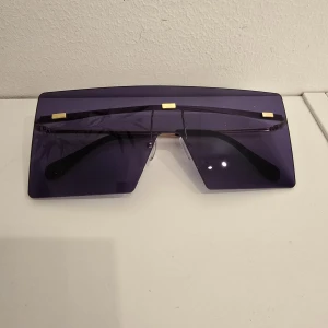 Lila futuristiska solglasögon - Unika solglasögon med stora, lila tonade sköldformade glas och guldiga detaljer upptill. Riktigt futuristisk vibe och perfekt accessoar för dig som vill sticka ut. Snygg och modern design med raka linjer och markerad form.