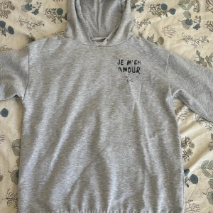 Grå hoodie från Gina Tricot med tryck - Grå hoodie från Gina Tricot med svart texttryck 'JE M'EN AMOUR' och en stjärna på bröstet. Mjuk insida och klassisk huva. Perfekt för dig som gillar en chill och enkel stil.