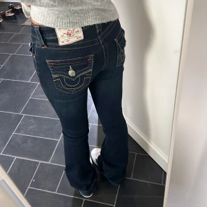 True Religion mörkblå bootcut jeans - Säljer ett par mörkblå jeans från True Religion med bootcut passform. Jeansen har snygga kontrastsömmar, stora fickor med lock och silverfärgade knappar bak. Klassisk logga på bakfickan och coola detaljer som sticker ut. Perfekta för dig som gillar street och premium denim.