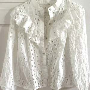 Vit blus med volang och hålbroderi - Superfin vit blus i bomull med broderade hålmönster och volangdetaljer över axlarna. Långärmad modell med knappar framtill❤️ Endast använd ett par gånger. Storlek XS men passar även S
