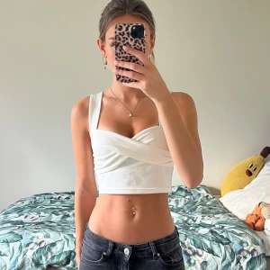 Vit croppad omlottopp med breda axelband - Säljer en vit croppad topp med breda axelband och snygg omlottdesign framtill. Toppen är ärmlös och har en tight passform som framhäver midjan. Materialet är stretchigt, troligen bomull eller polyester. 