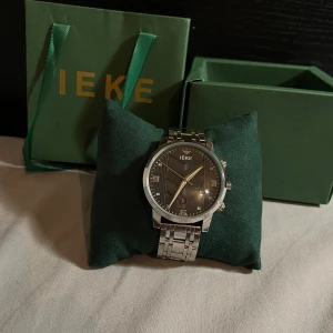 IEKE silverfärgad klocka med svart urtavla - Stilren klocka från IEKE med silverfärgad länkarmband i metall och svart urtavla med diskret mönster. Klockan har tydliga siffror, datumvisning! Den är helt oanvänd och funkar perfekt!  Vid intresse är de bara att kontakta!🙂