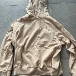 Beige hoodie med huva och ficka - Säljer en stilren beige hoodie i mjuk bomull med huva och dragsnören. Tröjan har en klassisk känguruficka framtill och ribbade muddar vid ärmslut och nederkant. Perfekt för en chill och avslappnad look.