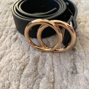 Svart bälte med dubbel ring-spänne - Stilrent svart bälte med snyggt dubbel ring-spänne i guld. Bältet är tillverkat i skinn och passar perfekt till jeans eller kjol. Enkel design som lyfter vilken outfit som helst med sin coola detalj framtill.