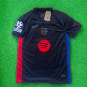 FC Barcelona Lamine Yamal tröja Nike - Snygg FC Barcelona matchtröja från Nike med Lamine Yamal och nummer 19 på ryggen. Tröjan är svart med röda och blå detaljer, har korta ärmar och klubbmärke samt sponsorer som Spotify och UNHCR tryckta. Perfekt för dig som älskar Barca och vill sticka ut på läktaren.