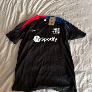FC Barcelona svart Nike fotbollströja S - Svart FC Barcelona fotbollströja från Nike med vita loggor och sponsortryck. Tröjan har korta ärmar och detaljer i rött och blått på axlarna. Materialet är lätt och andas, perfekt för träning eller match. Säljes tillsammans med matchande shorts.