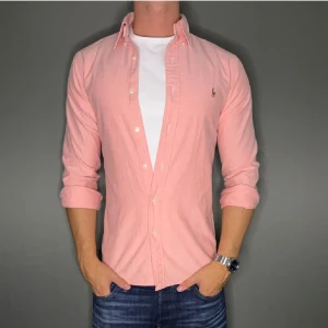 Ljusrosa skjorta från Ralph Lauren - Snygg ljusrosa skjorta från Ralph Lauren med klassisk krage och knappar hela vägen. Skjortan har lång ärm och ikonisk broderad logga på bröstet. Tillverkad i mjuk bomull och passar perfekt till jeans för en clean look.